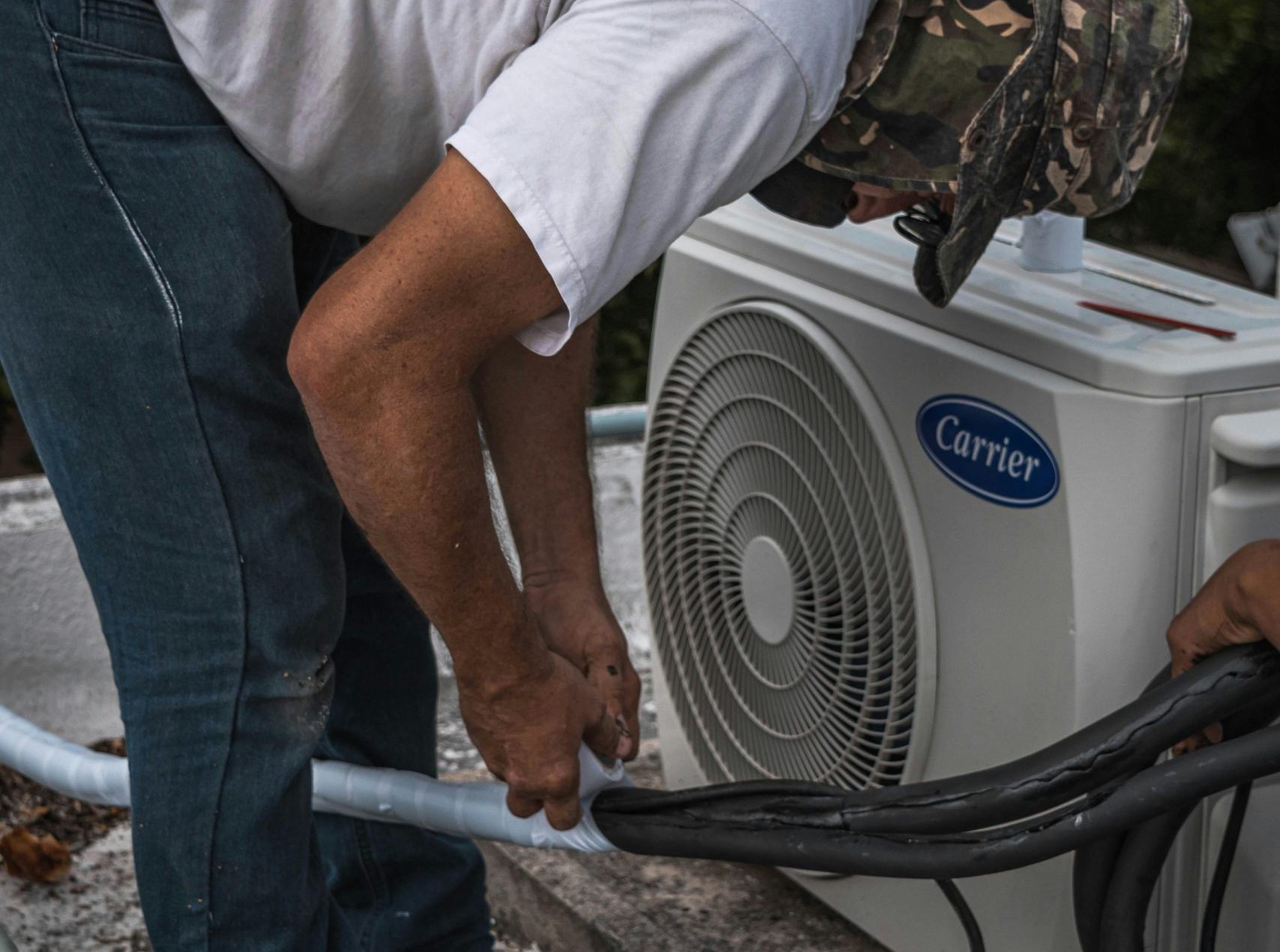 A technician performs maintenance on an outdoor air conditioning unit, focusing on hose connections.