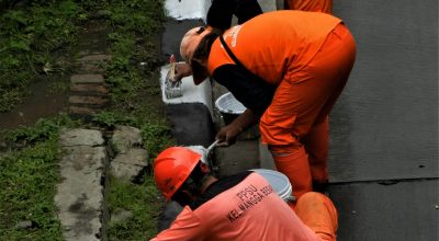 Three workers painting a curb in Jakarta, wearing orange uniforms, in a street maintenance task.